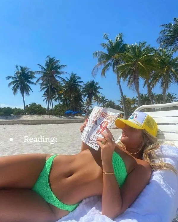  A woman in a bikini is laying on a chair on the beach reading a book.