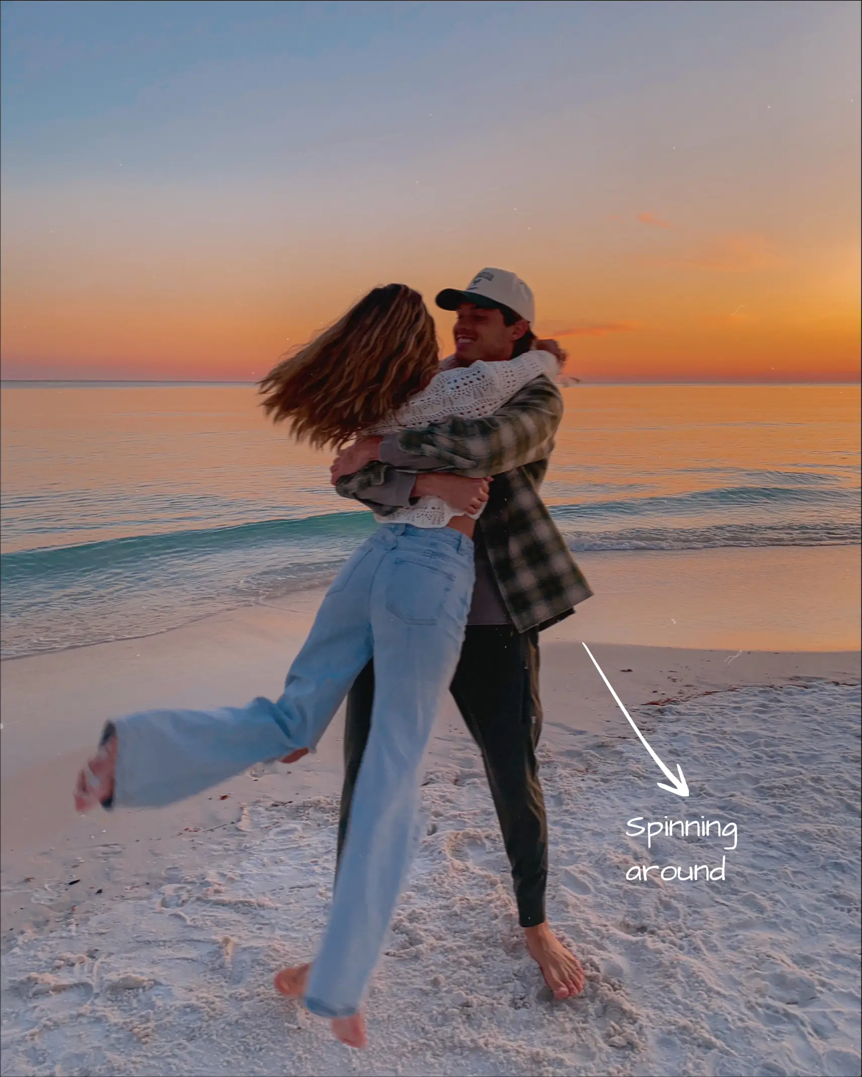  A man and a woman are hugging each other on a beach. The man is wearing a hat and jeans, while the woman is wearing a white shirt and jeans. The couple is spinning around each other, and they are both