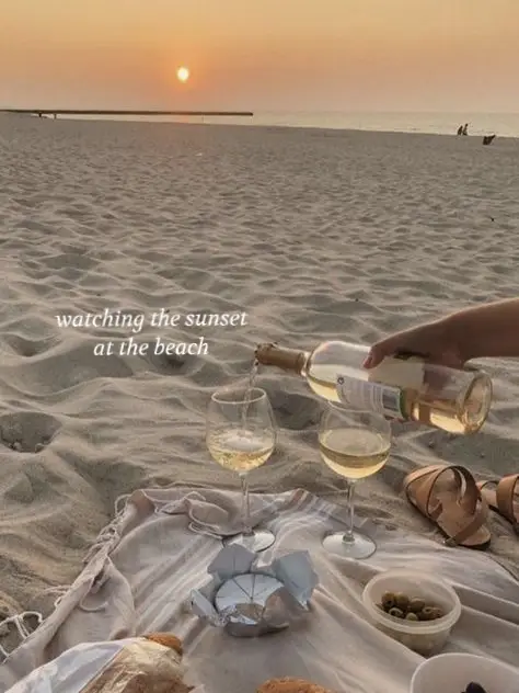  A person is sitting on a blanket at the beach, holding two wine glasses. The glasses are filled with wine, and the person is watching the sunset.