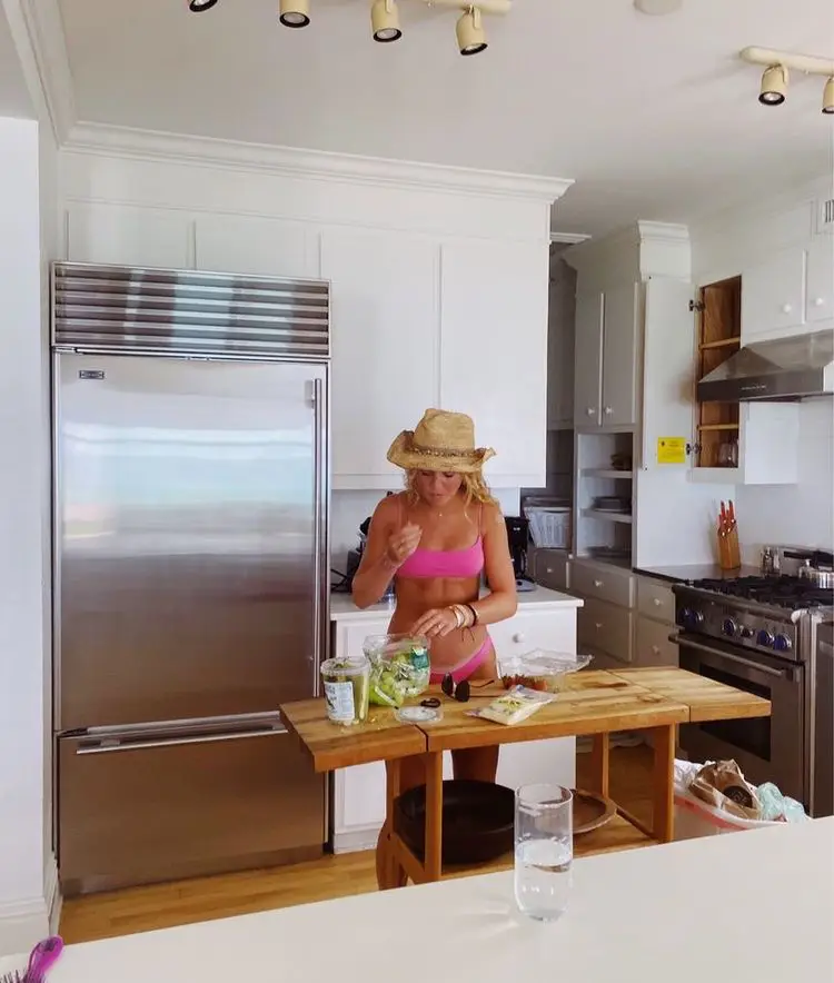  A woman is standing in a kitchen wearing a bikini and a hat. She is cutting up vegetables on a wooden cutting board.