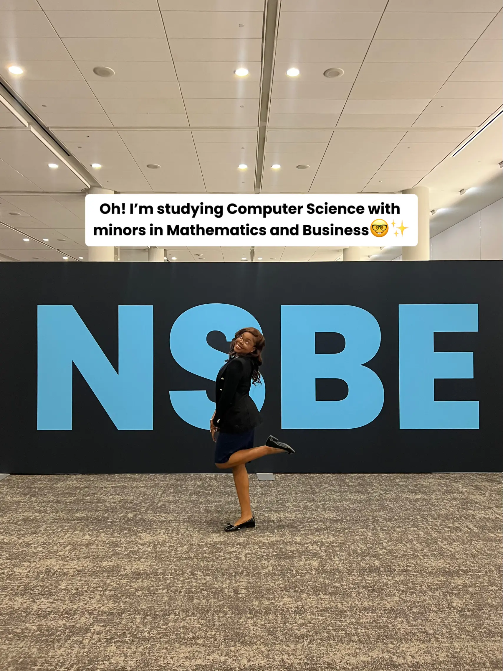  A woman is standing in front of a large sign that says "NSBE". She is wearing a black jacket and is smiling.