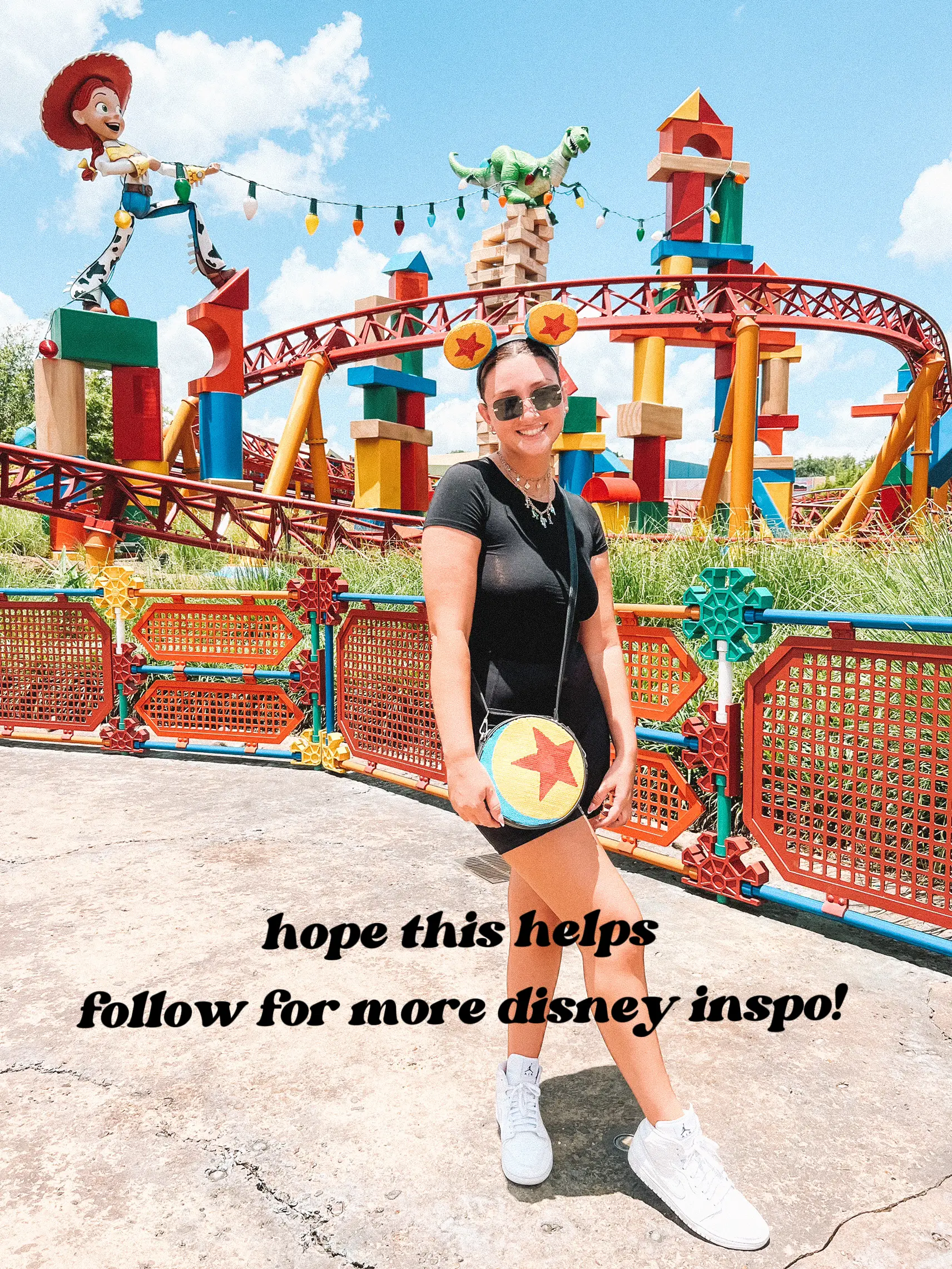  A woman wearing a black shirt and sunglasses is standing in front of a ferris wheel at a theme park. She is holding a frisbee and has a backpack on.