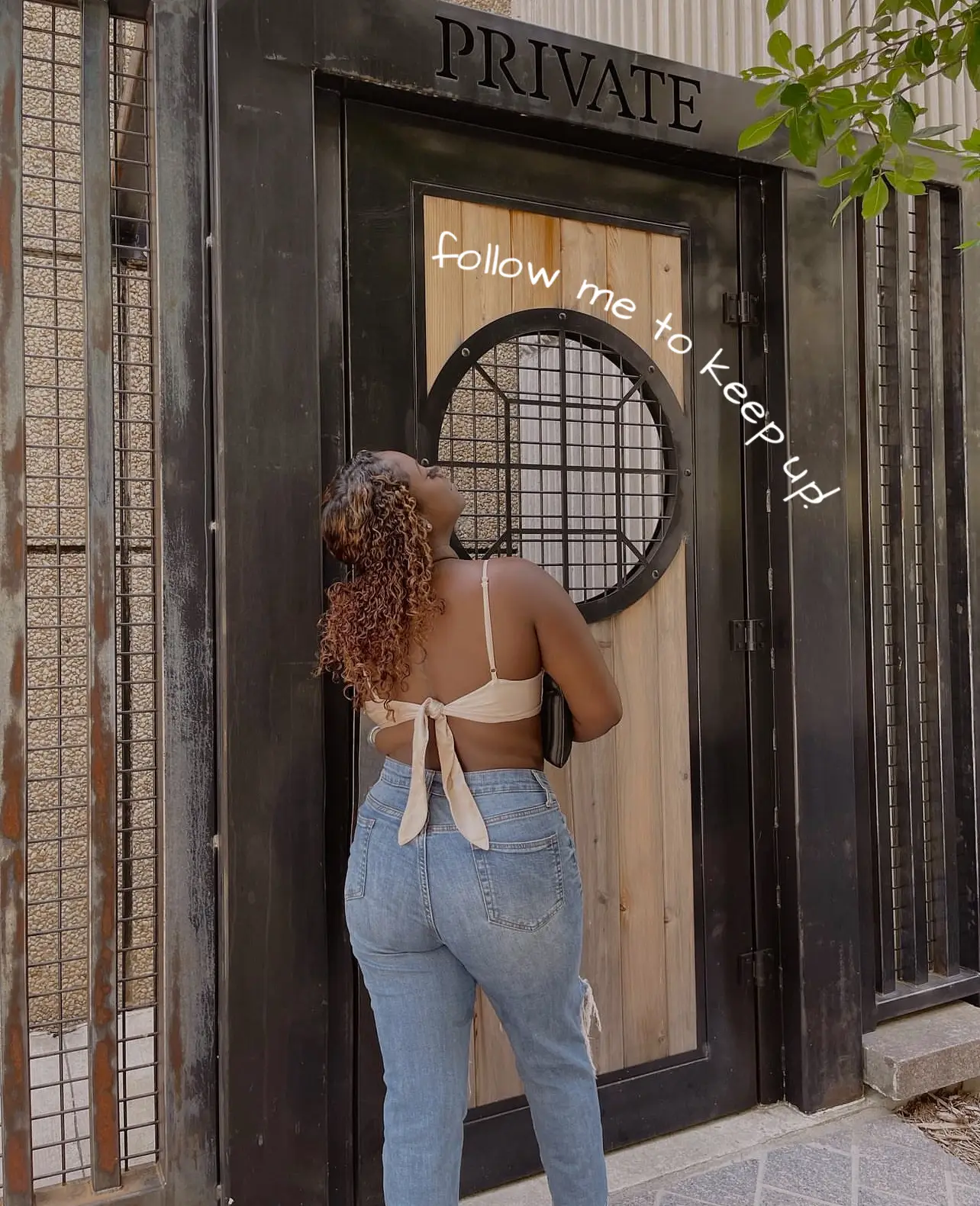  A woman is standing in front of a door, which has a sign above it that says "Private". The woman is wearing a white tank top and jeans.