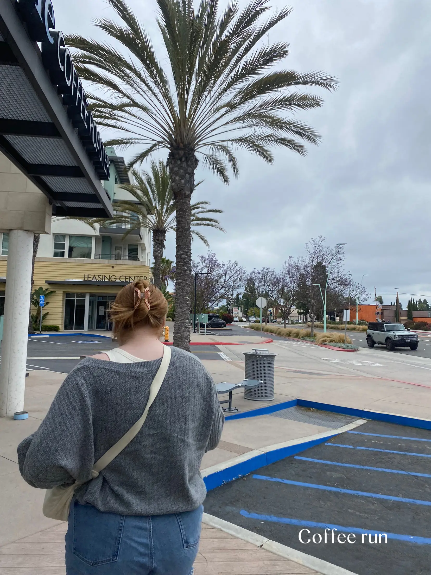  A woman is standing in a parking lot next to a building. She is wearing a brown sweater and jeans. The parking lot is empty and has a few cars parked in it. The woman is holding a coffee cup and a