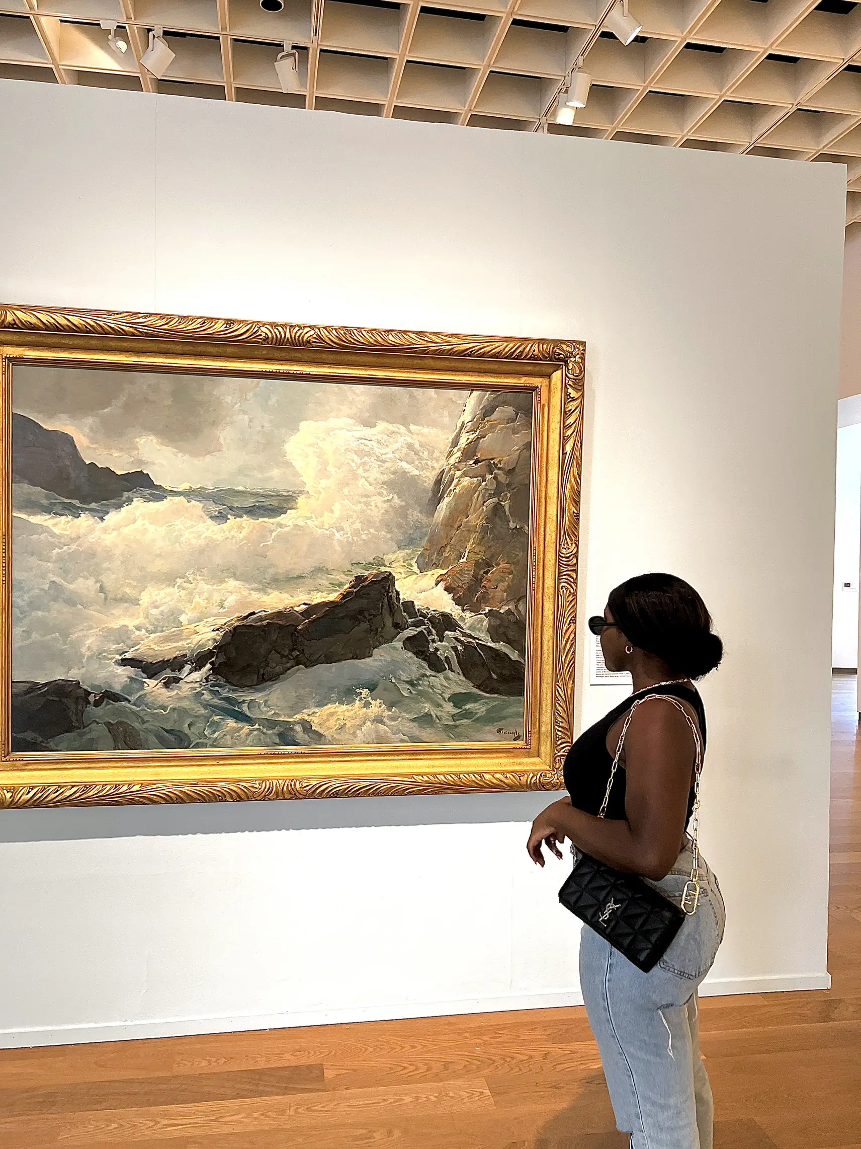  A woman in a black shirt and jeans is standing in front of a large painting. She is holding a purse and wearing a hat. The painting depicts a beach scene with a mountain in the background.