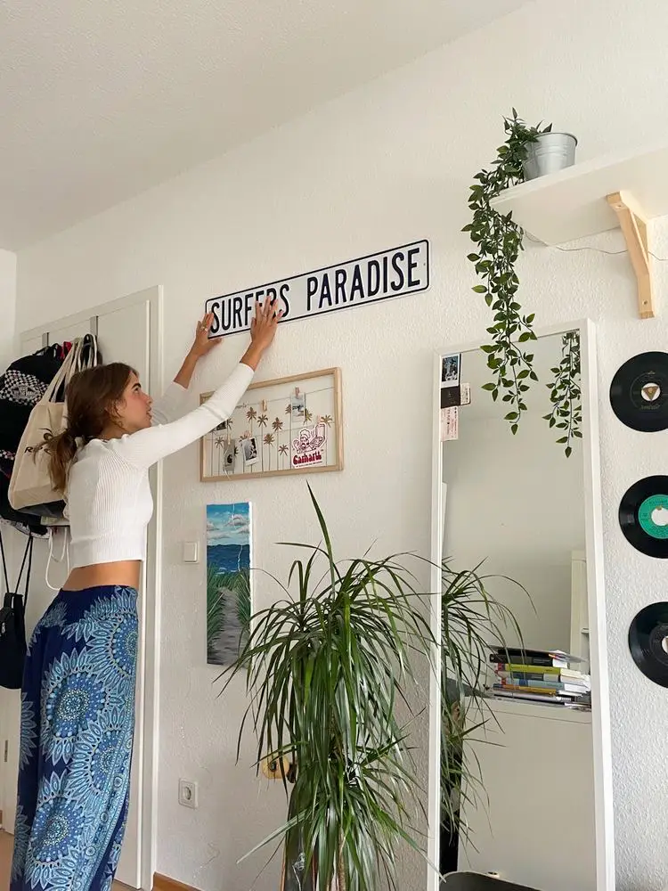  A woman is standing in a room with a sign that says "Surfers' Paradise". She is leaning against the sign and holding a potted plant. The room also has a bookshelf with books on it.