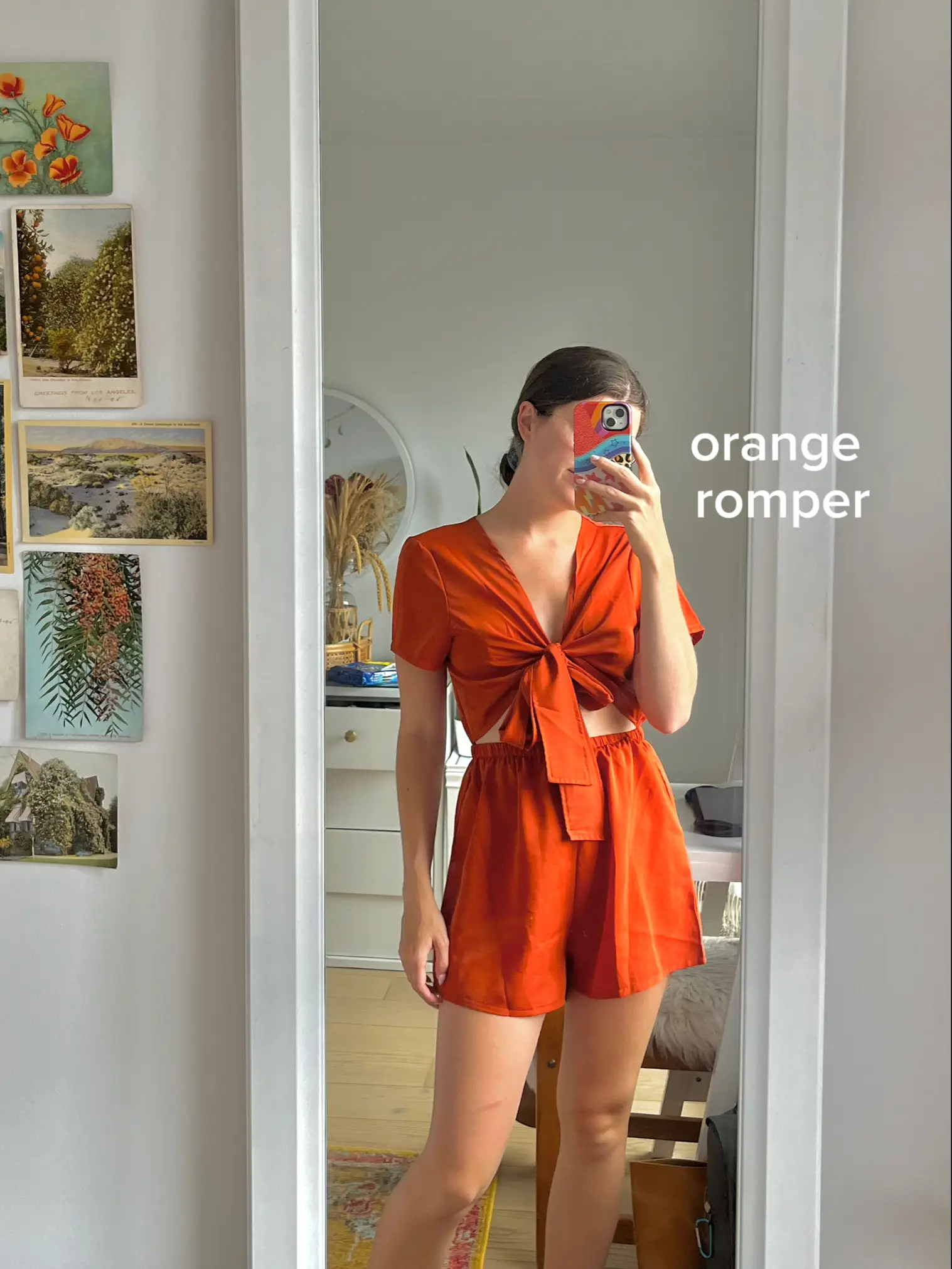  A woman in an orange romper is taking a selfie in a mirror.