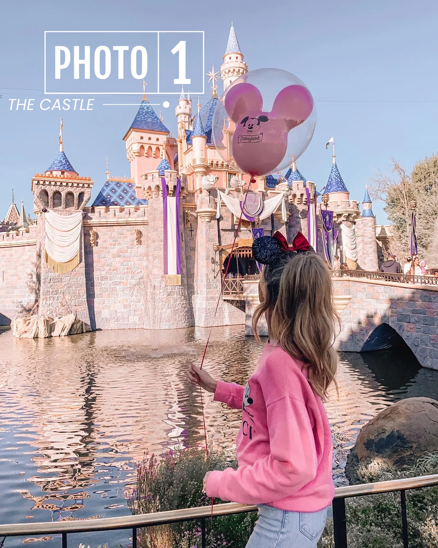  A little girl in a pink shirt is standing in front of a castle. She is holding a balloon and looking up.