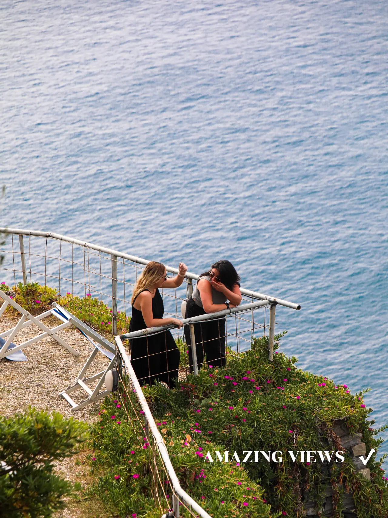 Two women are sitting on a railing overlooking the ocean.