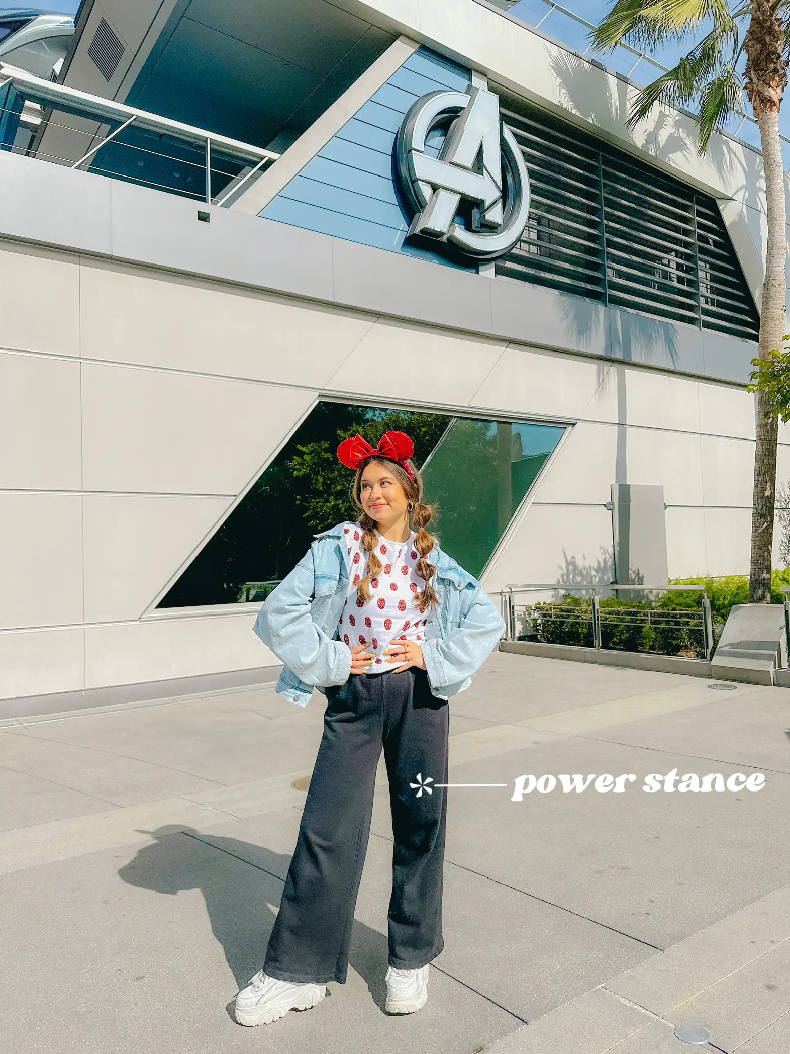  A woman wearing a blue shirt and jeans is standing in front of a building. She is wearing a Mickey Mouse headband and has her arms outstretched.