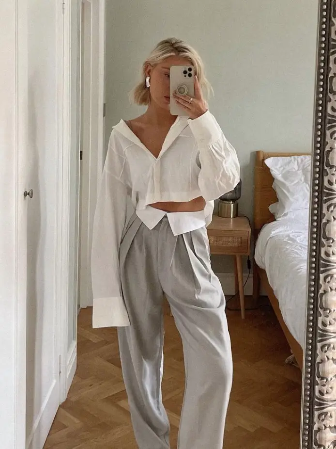  A woman wearing a white shirt and white pants is taking a selfie in a mirror.