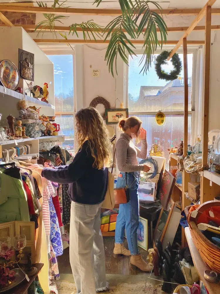  Two women are shopping in a store with a large display of ornaments. They are standing in front of the display, examining the items. There are several bottles and a vase on the display, as well as a few other people in the store.