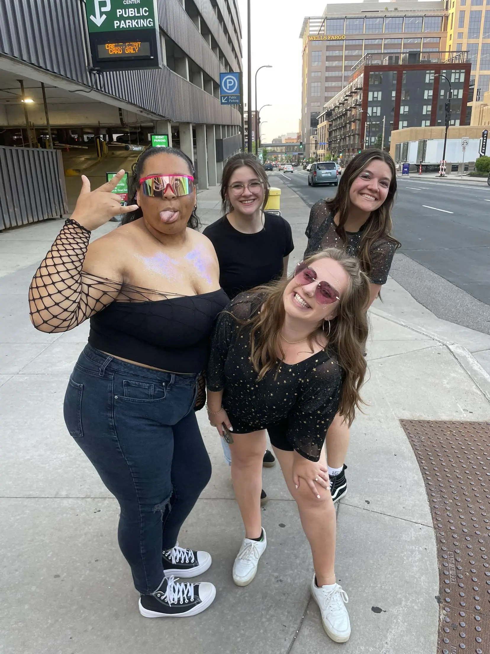  A group of women are standing on a sidewalk with their cars. They are posing for a picture and have their cars parked nearby. The women are wearing shades and jeans