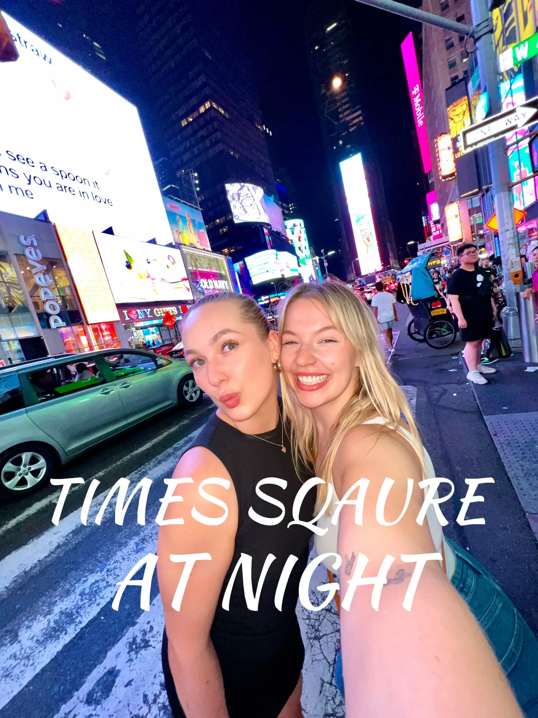  Two women are standing on a sidewalk in front of a building at night. They are posing for a picture and are smiling.