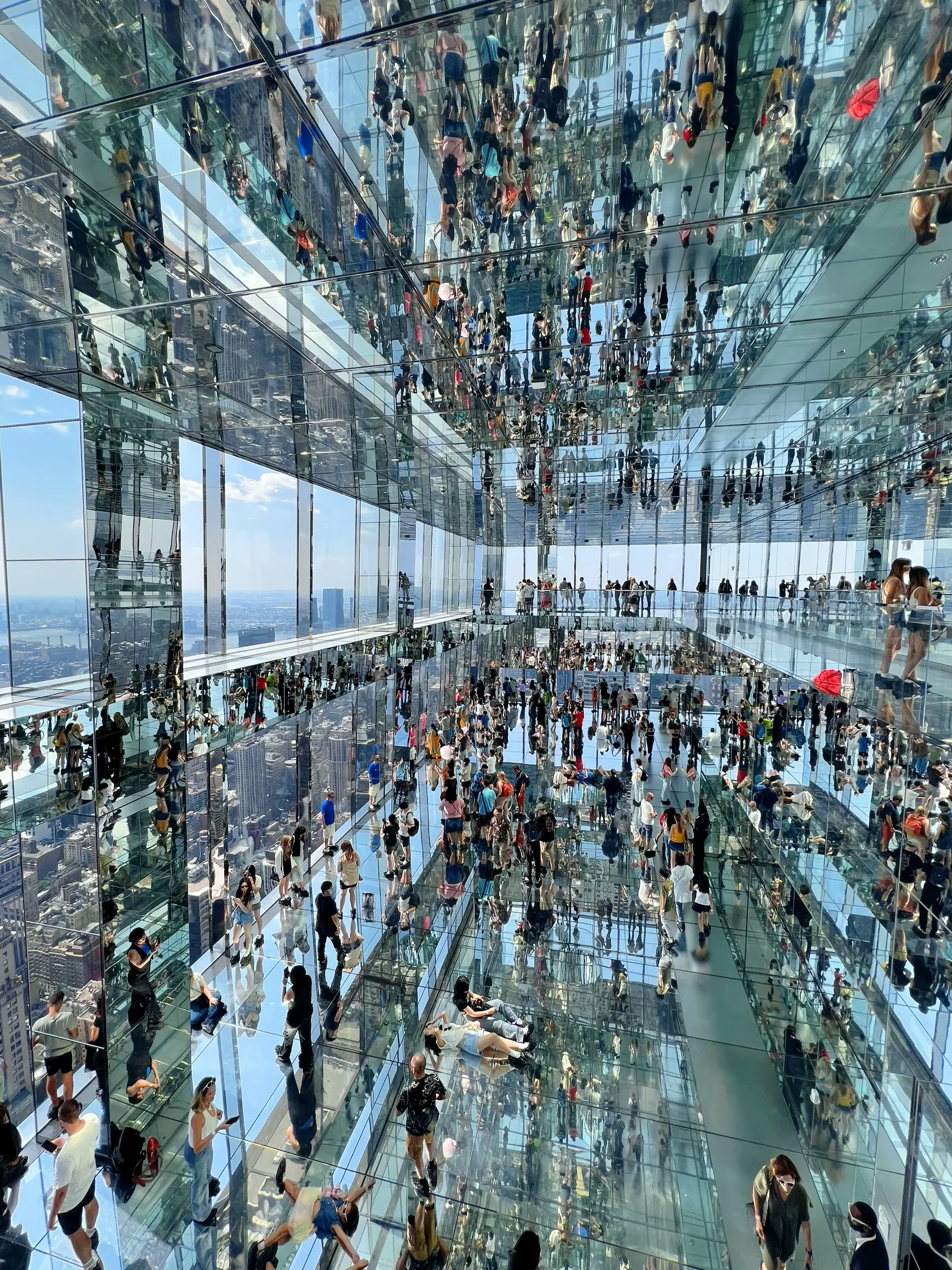  A large group of people are inside a building with a mirrored ceiling. The ceiling is filled with lights, and there are many people standing and walking around. Some people are holding umbrellas, possibly to protect themselves from the sun. The image is