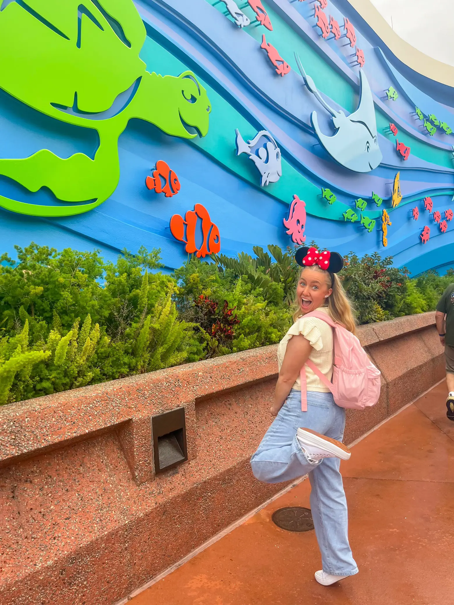  A woman wearing a white shirt and blue jeans is standing in front of a colorful fish wall. She is holding a frisbee and appears to be enjoying her time at the aquarium.
