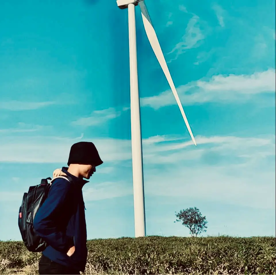  A man wearing a xanh rờn shirt and a hat is standing in front of a wind turbine.