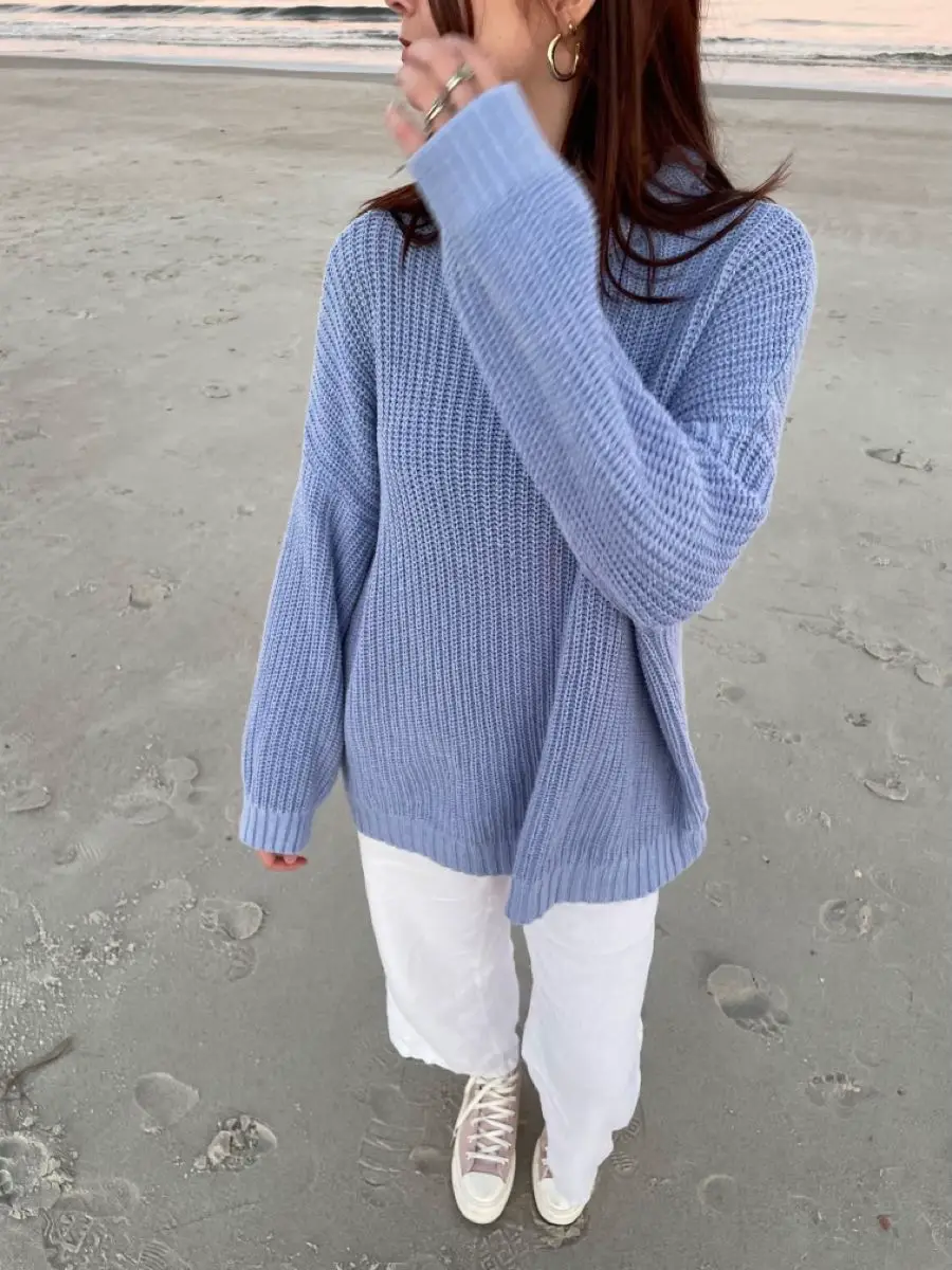  A woman wearing a blue sweater and white pants is standing on a beach.