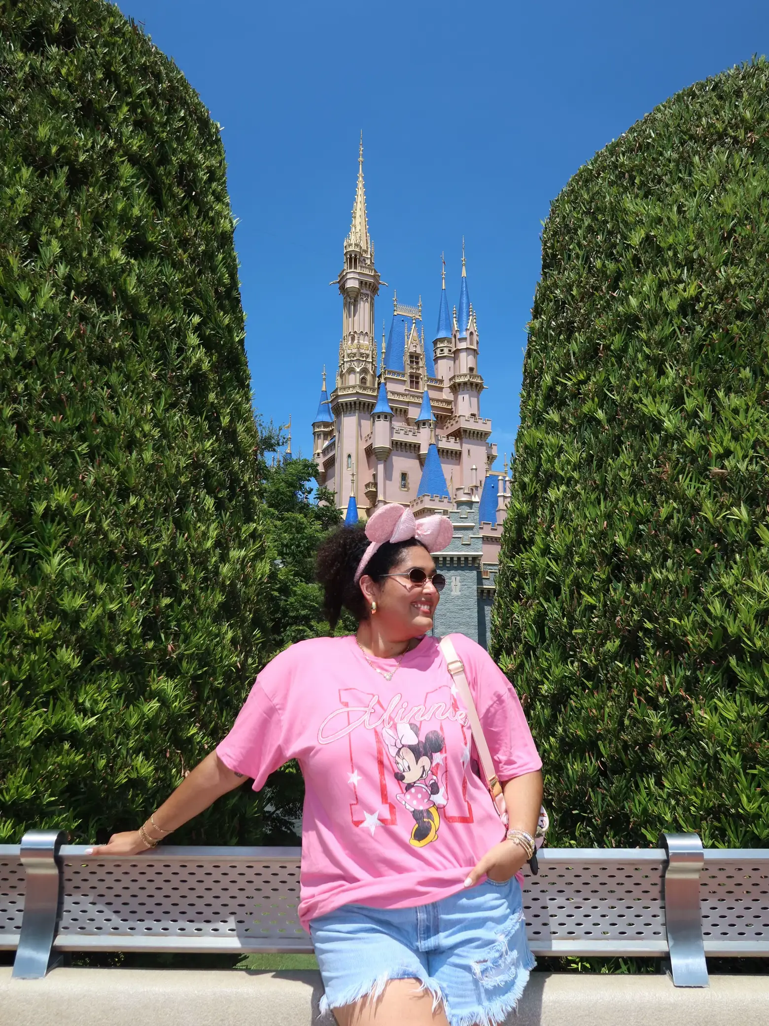  A woman wearing a pink shirt and a hat is sitting on a bench in front of a castle. She is smiling and holding a purse.