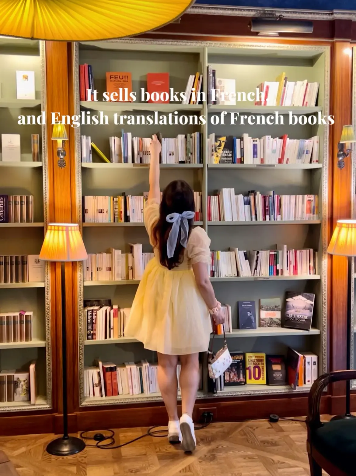  A young girl is standing in a bookshelf with a French flag on the wall. The shelf is filled with books in both English and French translations of French books.