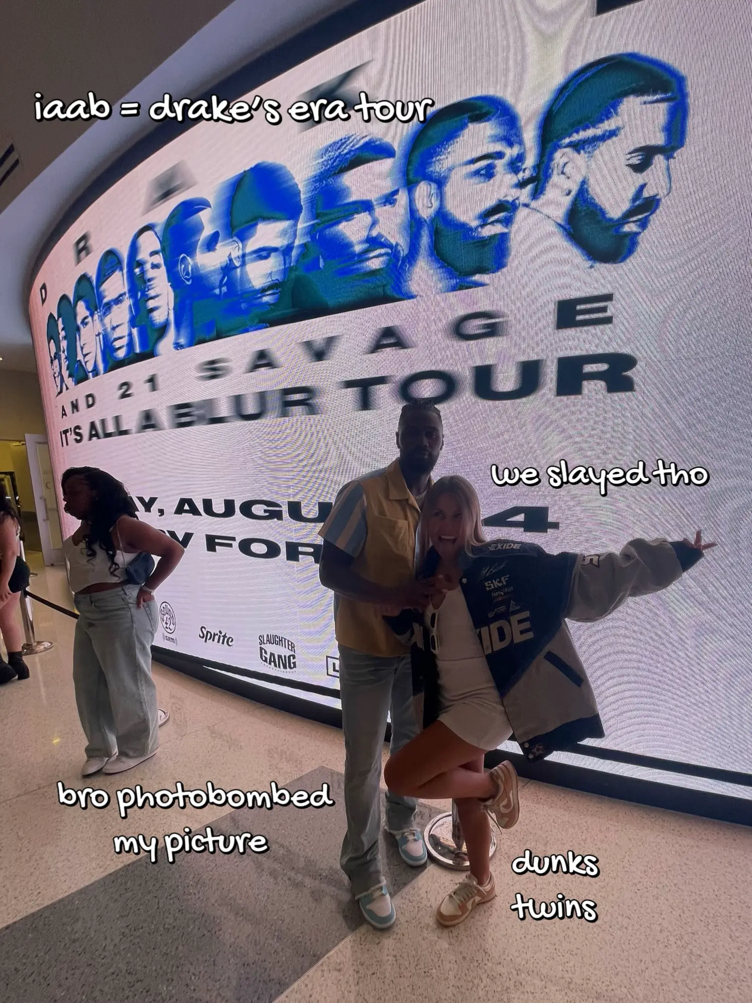  A man and a woman are standing in front of a large display of photos. The man is wearing a blue shirt and the woman is wearing a white shirt. The man is holding a cell phone and the woman is holding a cell phone. The display of photos includes people
