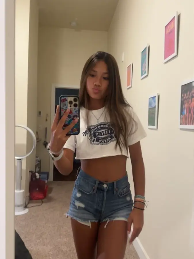  A woman in a white shirt and shorts is taking a selfie in a mirror.