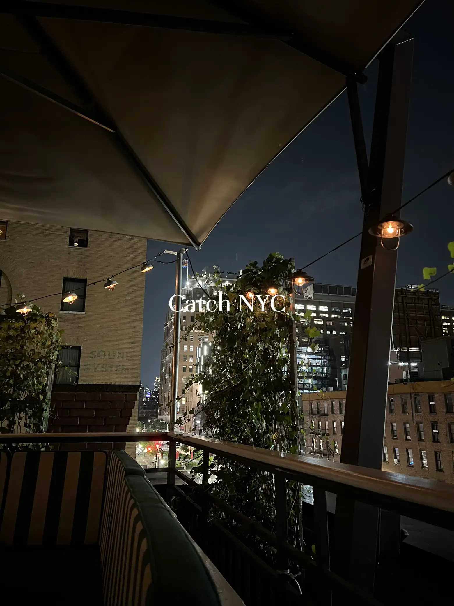  A view of a building at night with a patio and a tree. The patio has a striped couch and a chair. The building is lit up and has a sign that says "Catch NY".