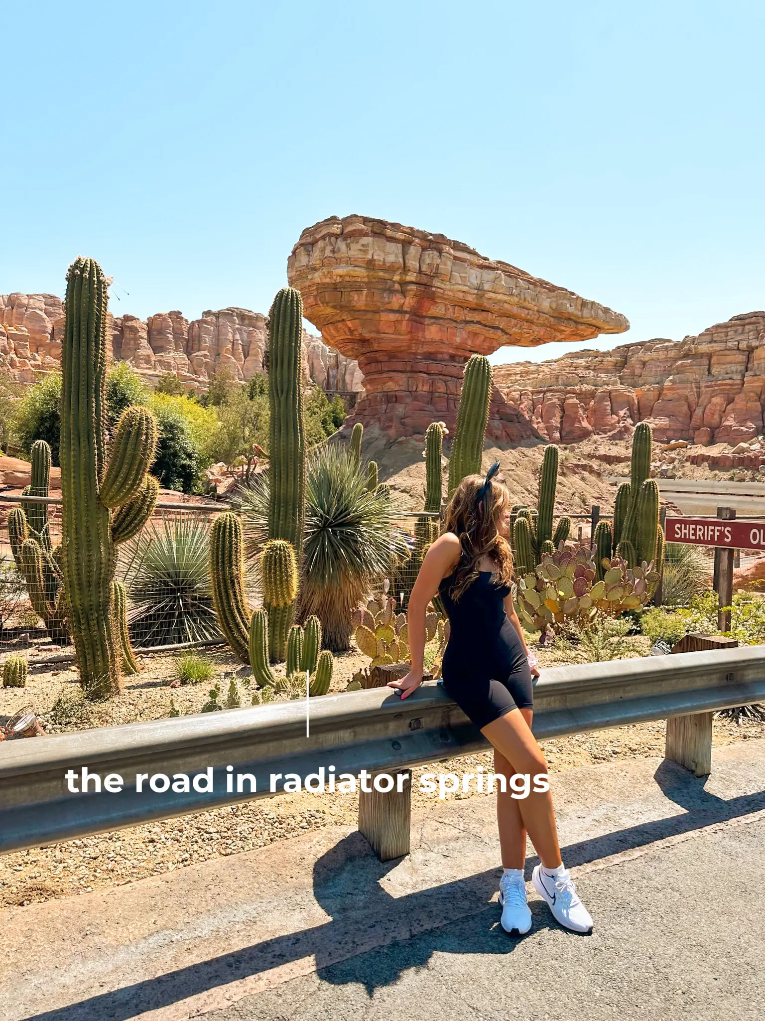  A woman is sitting on a fence near a road with cacti and a sign that says "road in radiator springs".