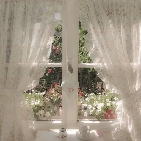  A white curtain with a window behind it. The curtain is white and has a Christmas tree on it. The window is filled with flowers and plants.