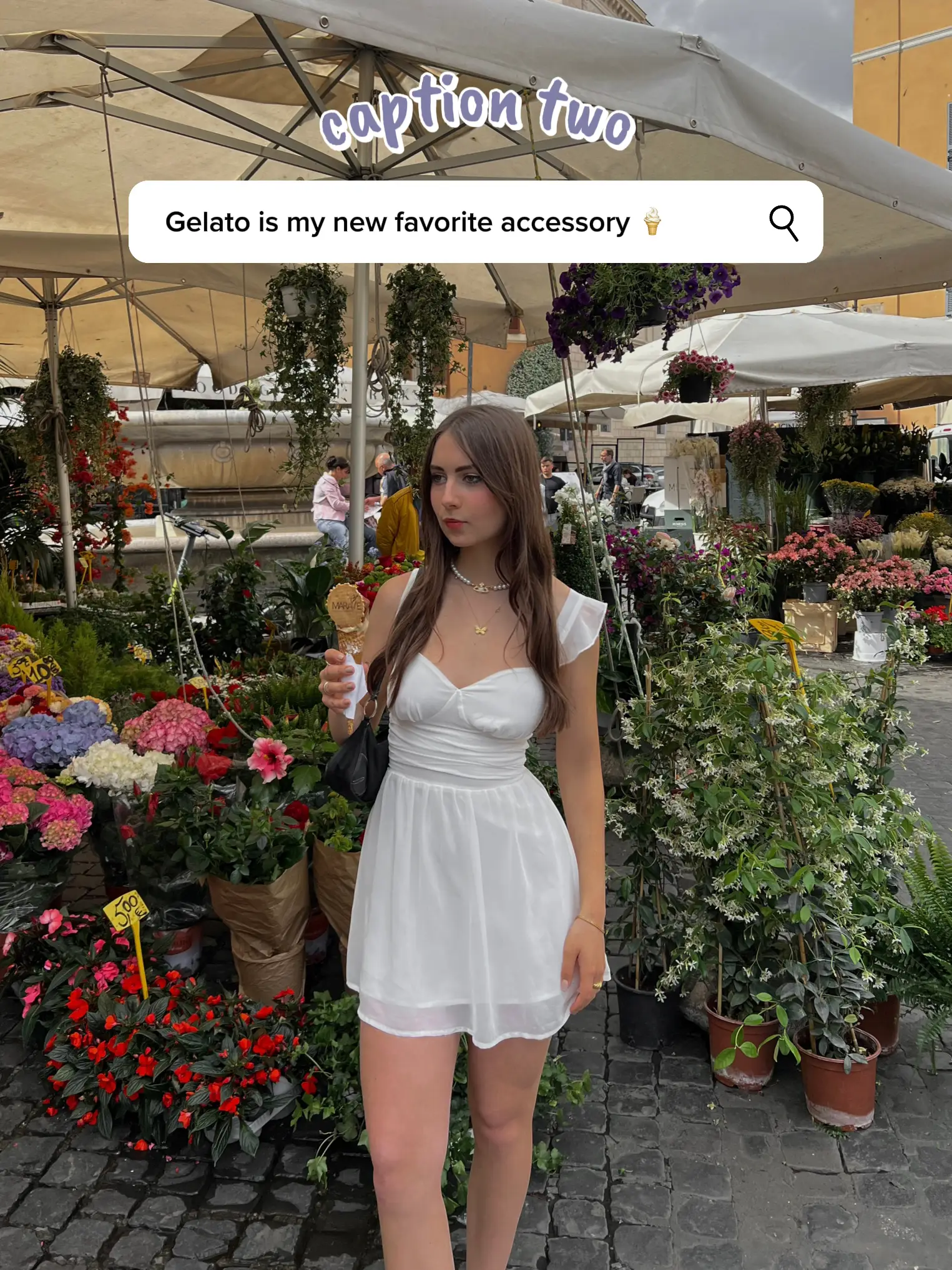  A woman in a white dress is standing in front of a flower shop. She is holding a glass of Gelato and looking at the camera. The shop is filled with flowers and vases, and there are