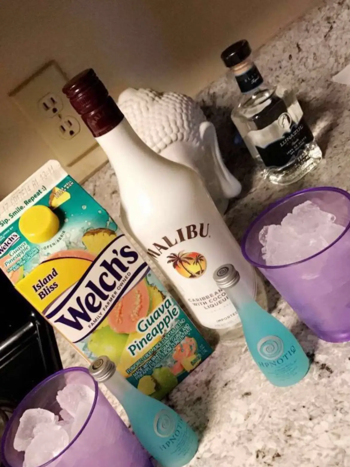  A bottle of Alibi is on a counter next to a cup of juice and a bottle of Welch's Bliss Island.