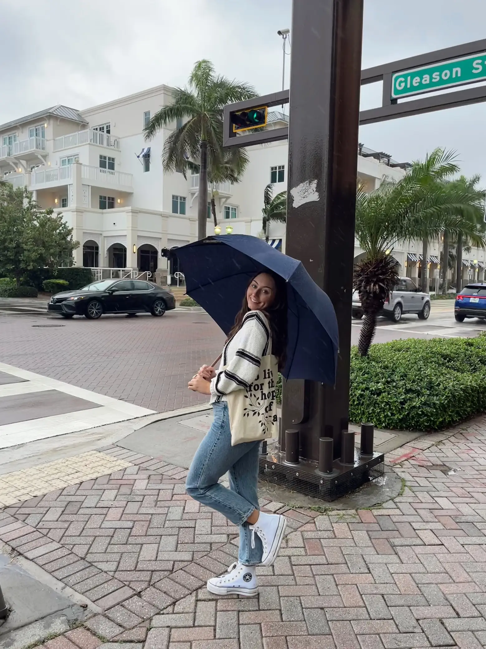  A woman is standing on a sidewalk holding an umbrella and a bag.