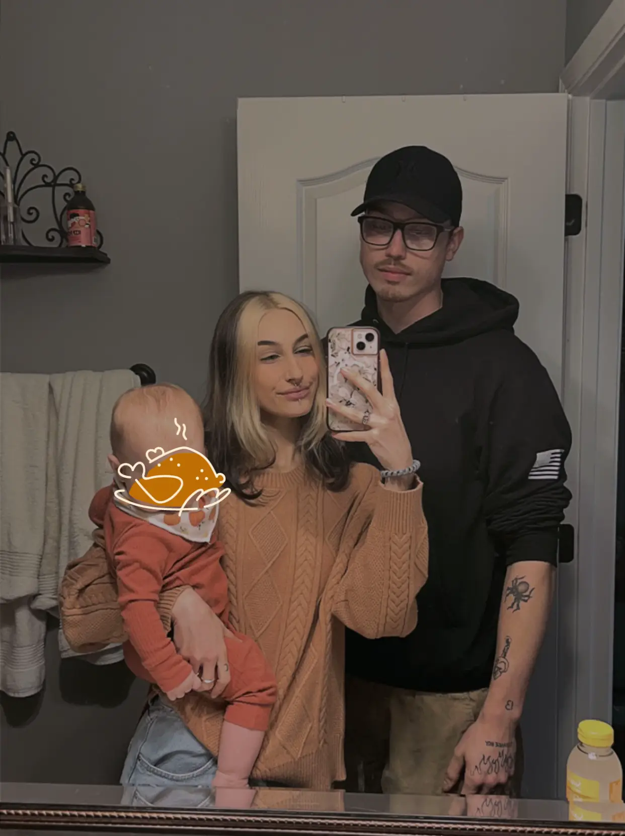  A man and a woman are standing in a bathroom. The woman is taking a selfie with the man and the baby. The man is wearing a black shirt.