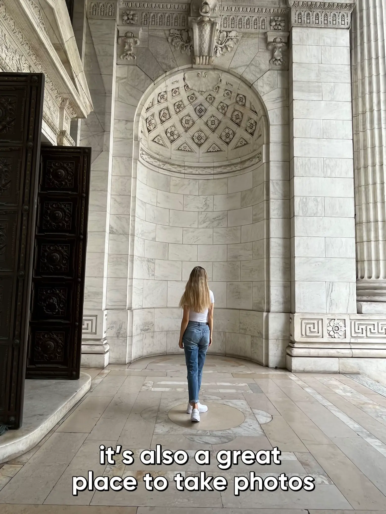  A woman is walking in front of a building with a archway. The building has a large window and a pillar. The woman is wearing a white shirt and jeans. The building is described as