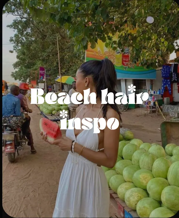  A woman in a dress is standing in front of a fruit stand with a sign that says "Beach Hairstyle". She is holding a watermelon and looking at the camera.