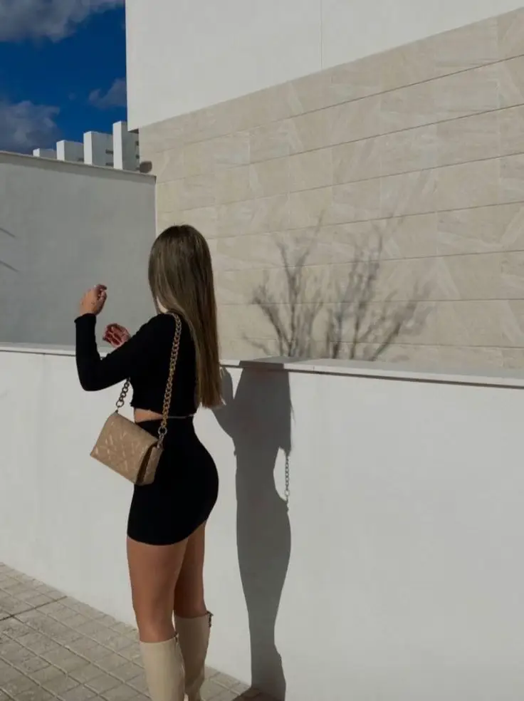  A woman in a đen giòn dress is standing in front of a white wall. She is holding a purse and looking up. The sun is casting a shadow on the wall, and the woman is standing in front of it.
