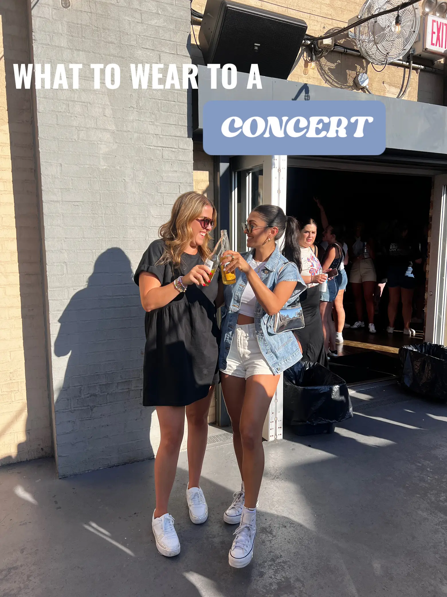  Two women are standing outside a building, wearing shirts and jeans. They are holding drinks in their hands, and there is a backpack nearby. The words "What to Wear to a Concert"