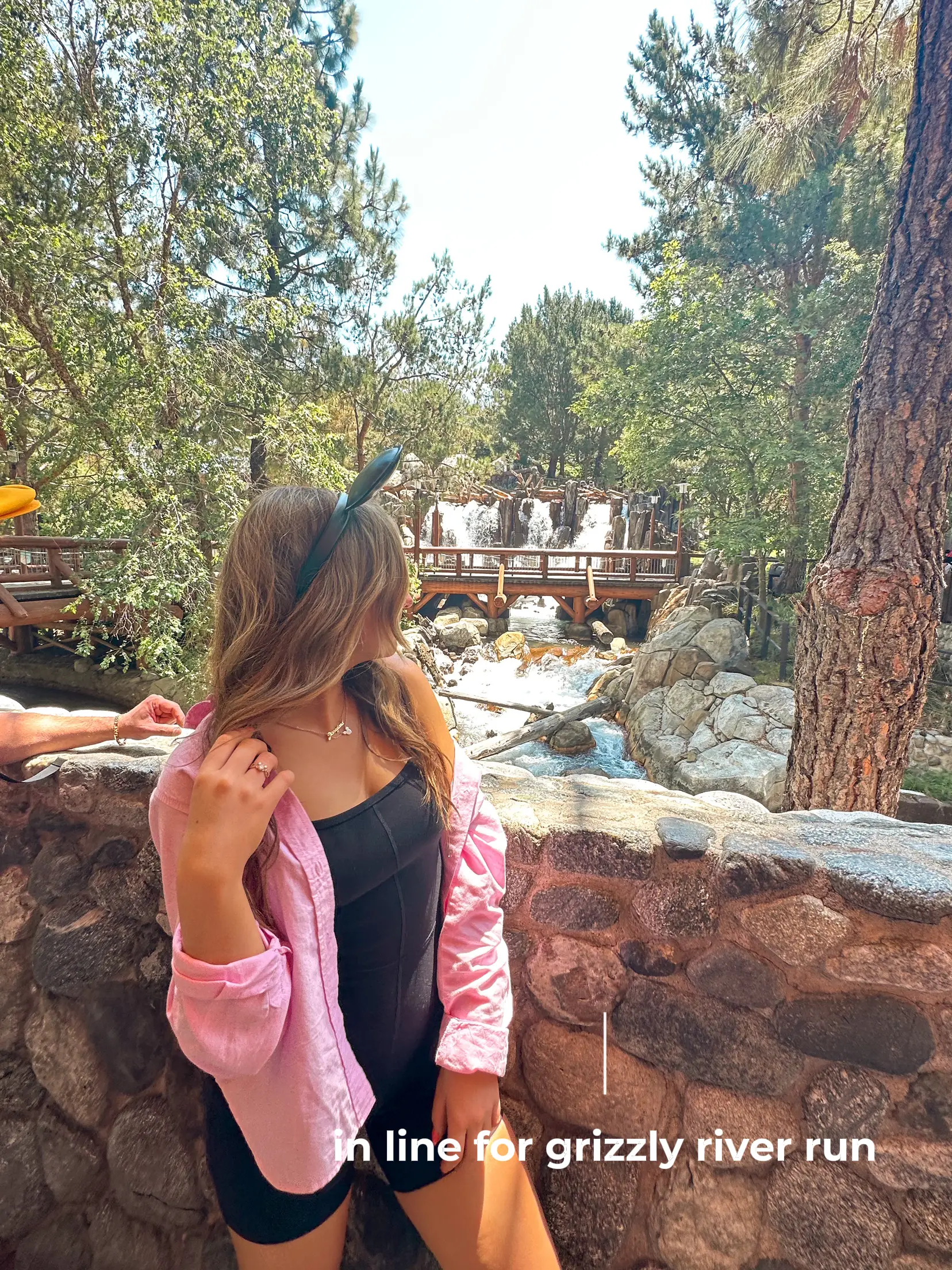  A woman wearing a pink jacket and black shorts is standing in front of a brick wall. She is wearing a hat and has a backpack on. The words "in line for grizzly river run"