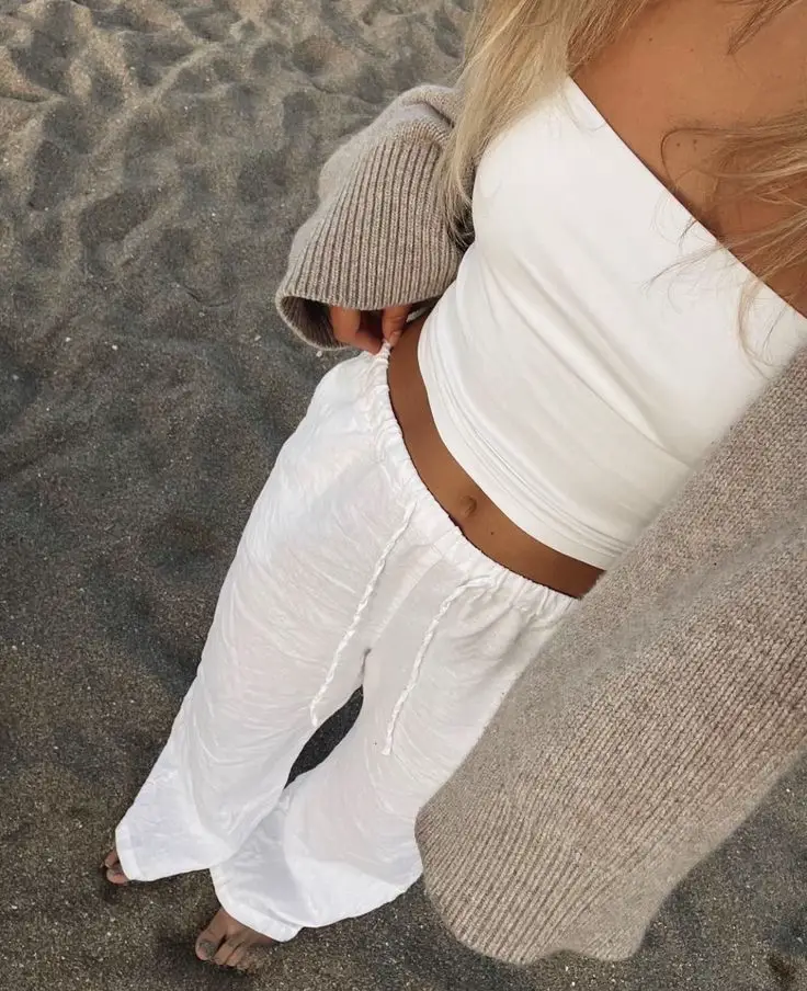 A woman wearing a white sweater and white pants is standing on a beach.
