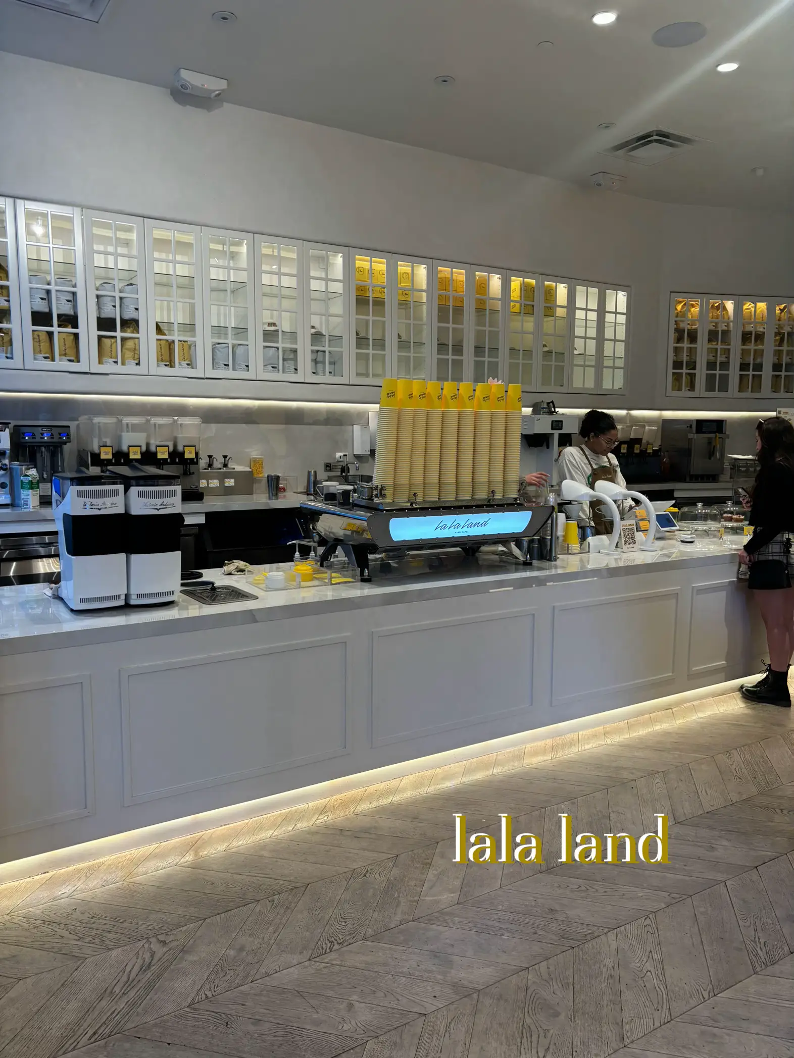  A restaurant with a man standing at the counter and a woman sitting at a table. The restaurant has a large menu board and a La Land sign.