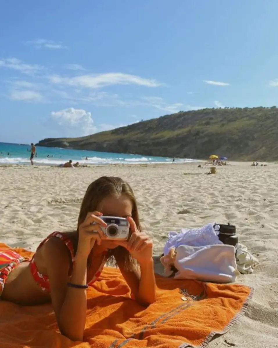  A girl is laying on a towel on the beach, taking a picture of the ocean.