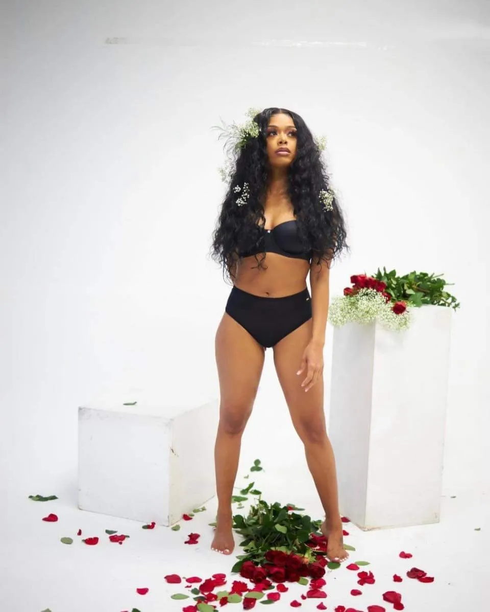  A woman wearing a black bikini top and black lace pants is standing in front of a wall. The wall has red roses on it.